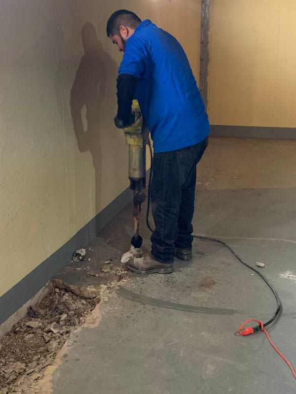 Man in blue shirt uses a jackhammer to break up concrete floor in a basement.