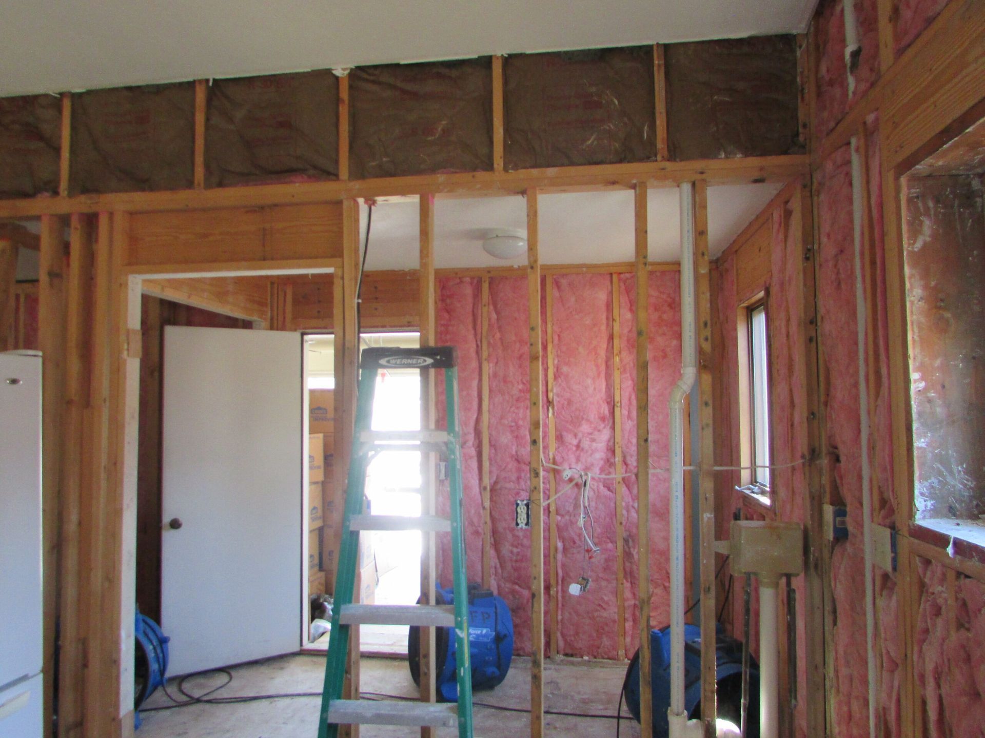 Interior view of a room under construction with exposed wooden framing and pink insulation. A ladder stands nearby.