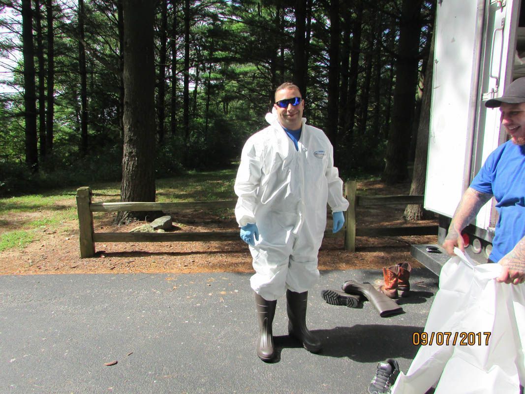 Man in white protective suit and boots, outdoors near forest, with a person holding a white bag.