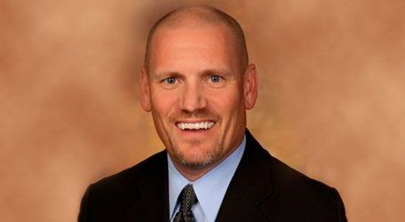 Headshot of a smiling man in a dark suit and tie against a warm tan background