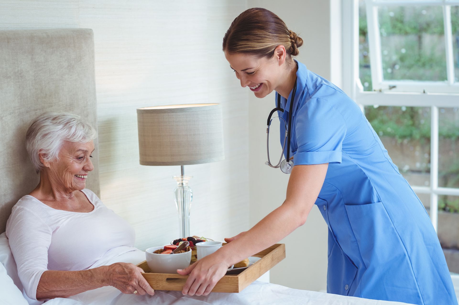 A nurse is serving food to an elderly woman in a hospital bed.
