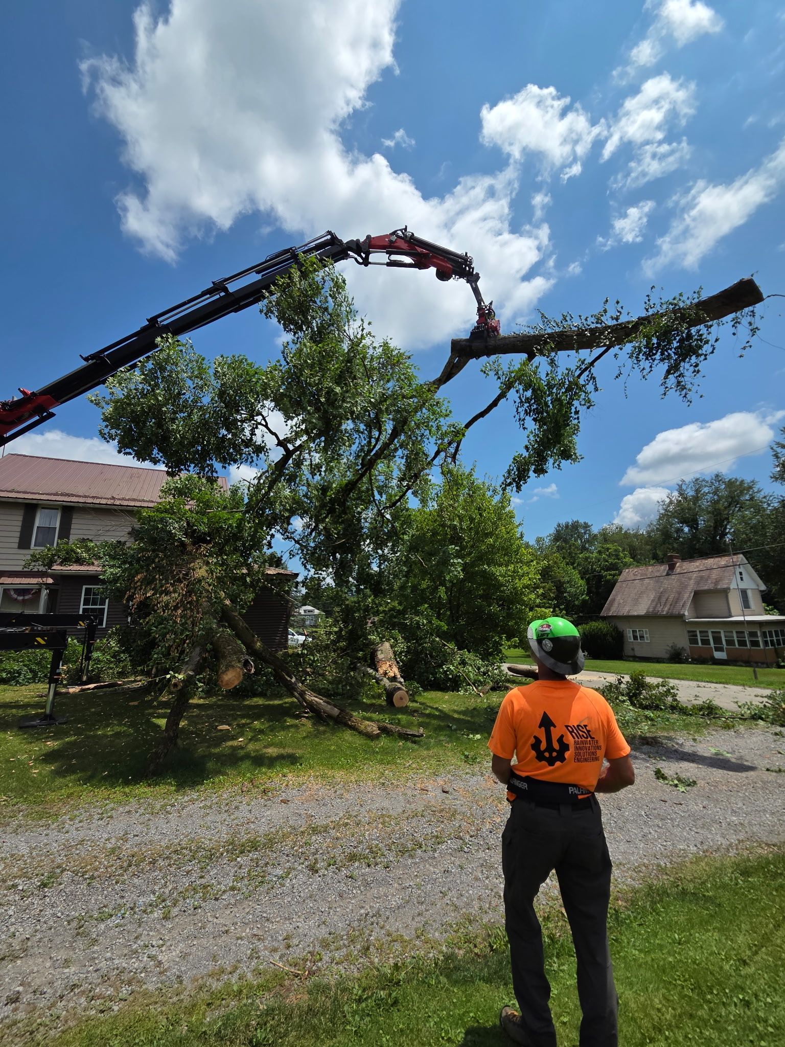 Worker using a crane trims a leaning tree beside a house on a sunny gravel driveway