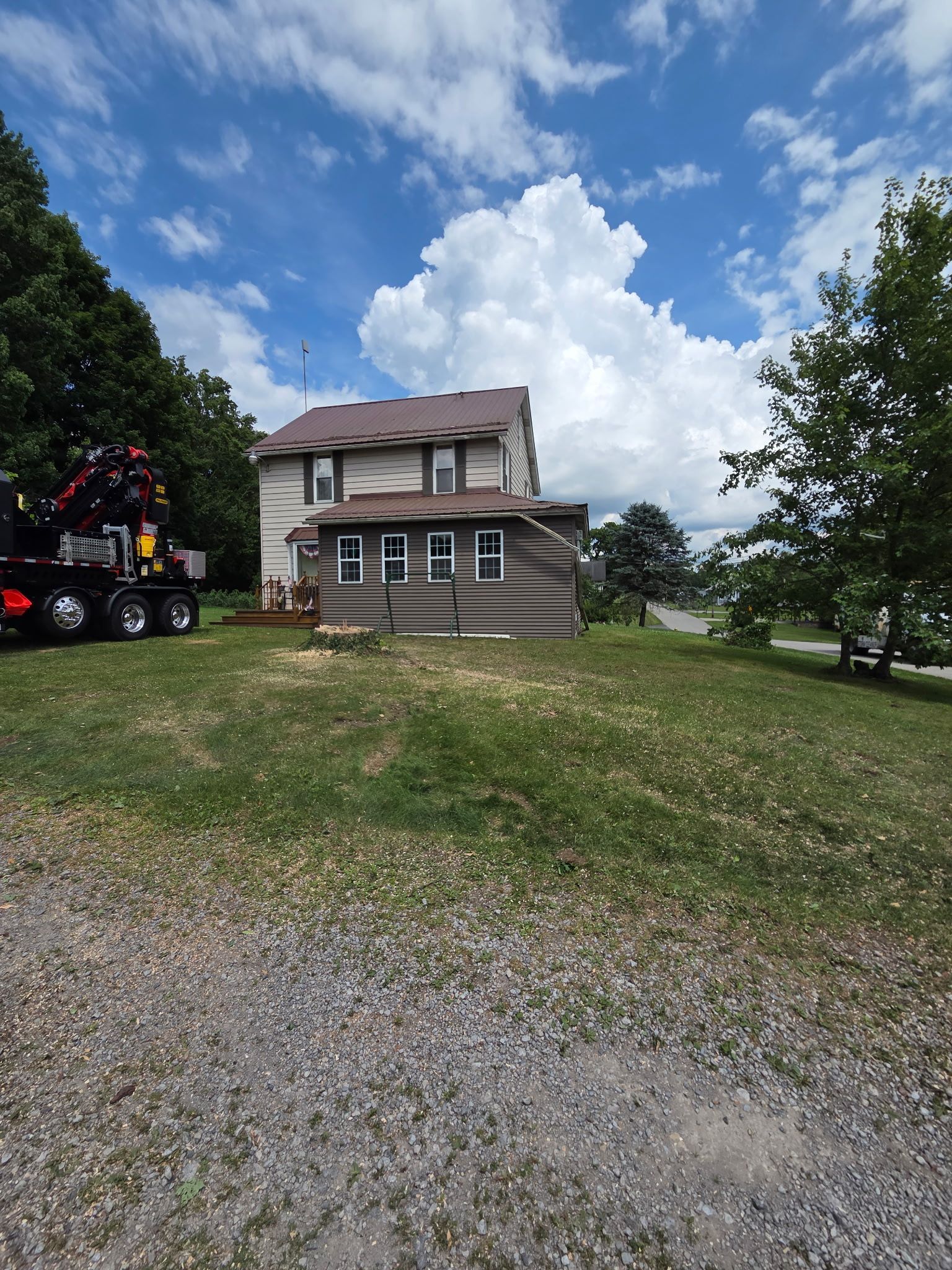 Rural two-story house with brown siding under a blue sky, with a truck and trailer parked on the left