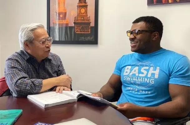 Two men at a table, looking at a book and talking. The man on the right wears a blue "BASH Swimming" shirt.