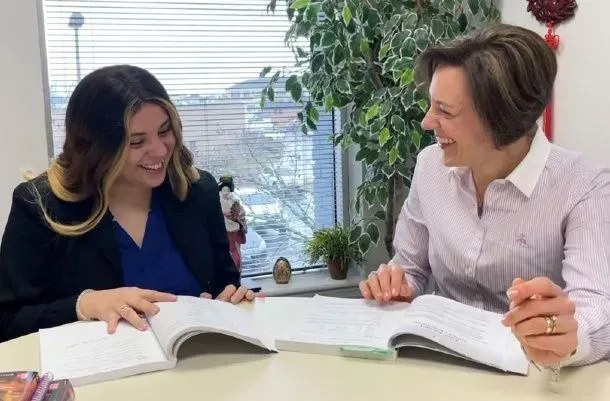 Two women smiling, sitting at a table with open books, near a window with a view.
