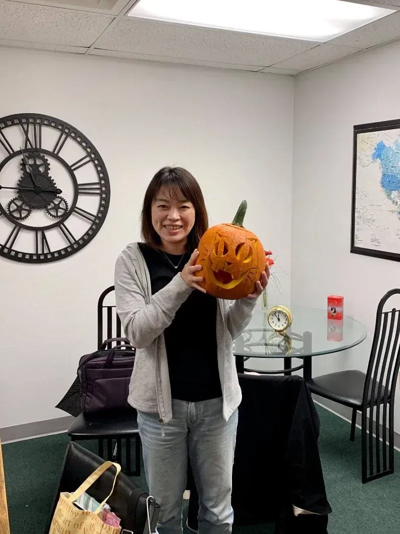 Woman holding a carved pumpkin with a smiling face in an office setting.