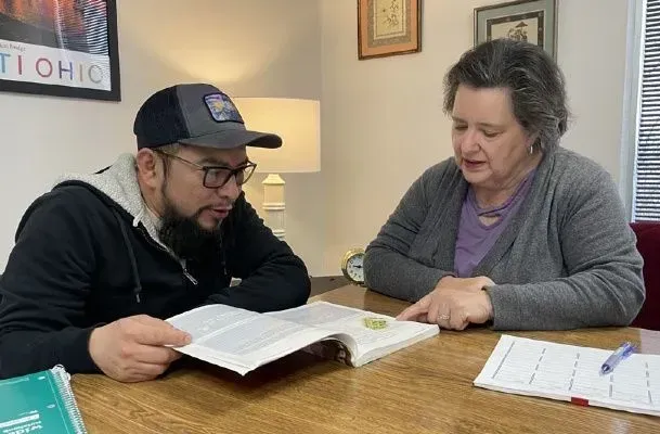 Man in hat and woman look at an open book, pointing. They sit at a table in a room.