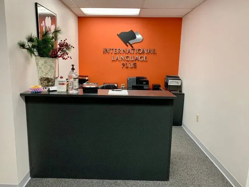 Reception desk at International Language Plus, orange wall with logo, black desk, printer, floral arrangement.