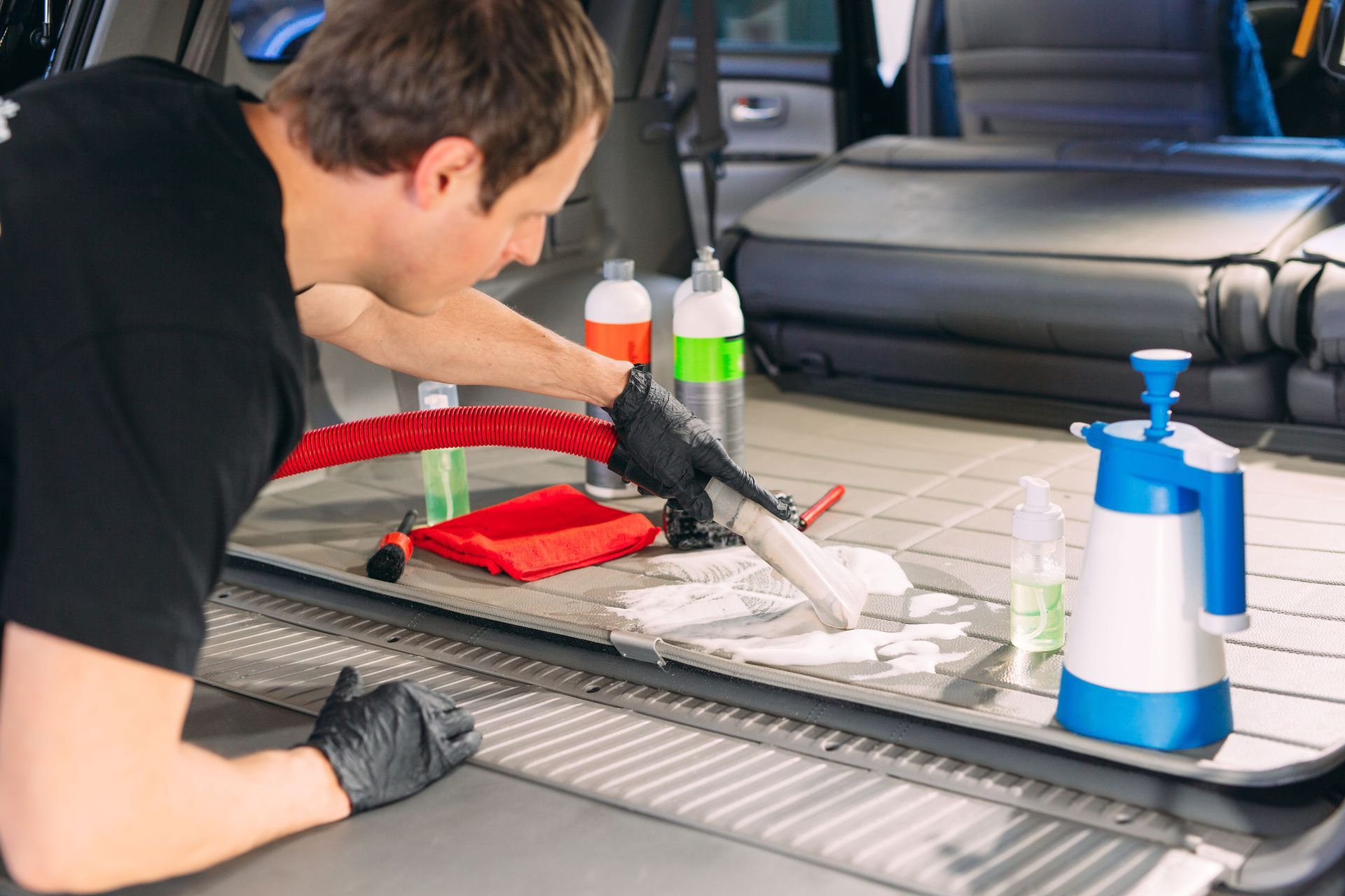 Man cleaning car trunk with tools and products, inside the car, wearing gloves.
