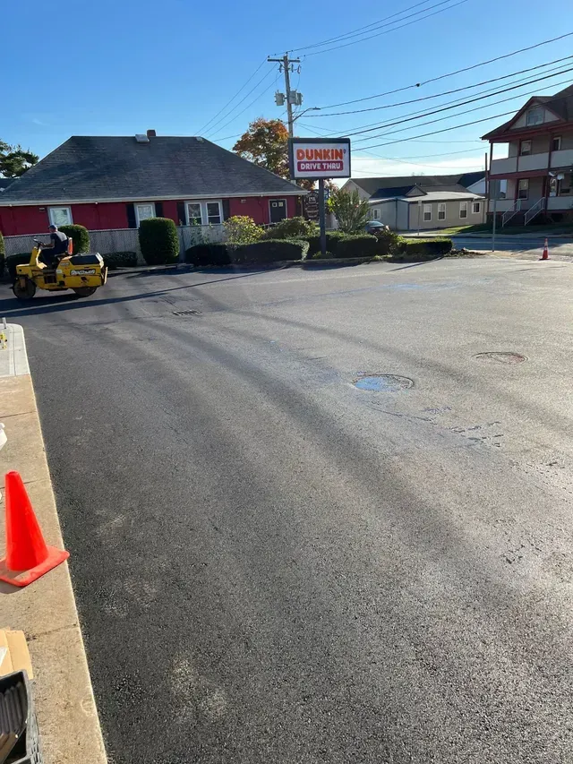 Freshly paved street outside a Dunkin' sign, with a roller and orange cone in the foreground.