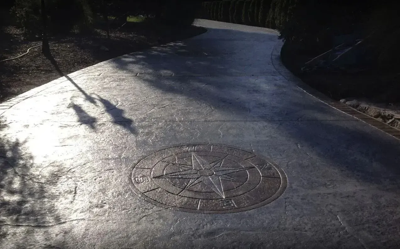 Stamped concrete driveway with a central compass rose medallion design, illuminated by sunlight and cast tree shadows.