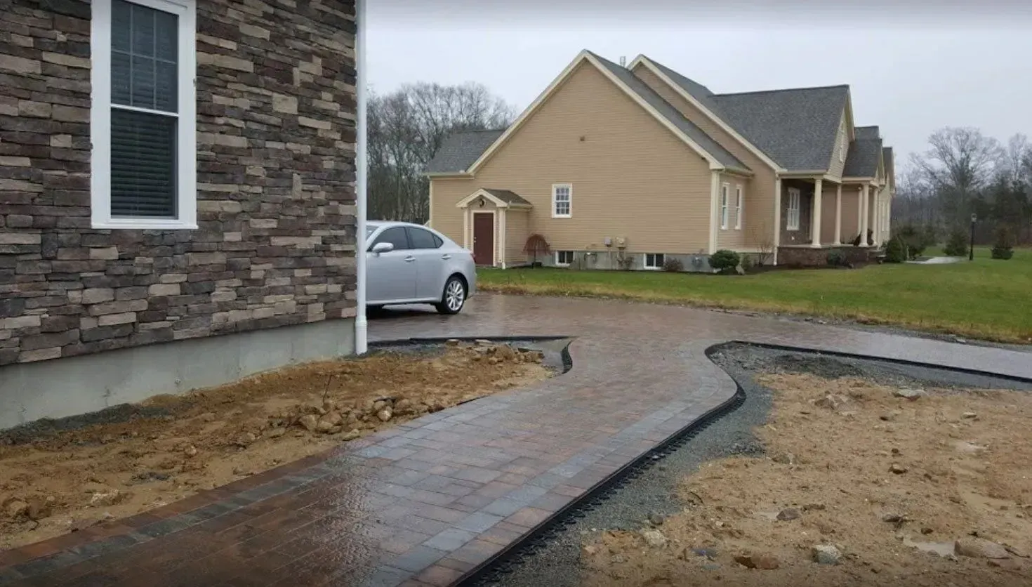 A paved walkway curves in front of a stone house, leading toward a tan house and a silver sedan in a suburban yard.
