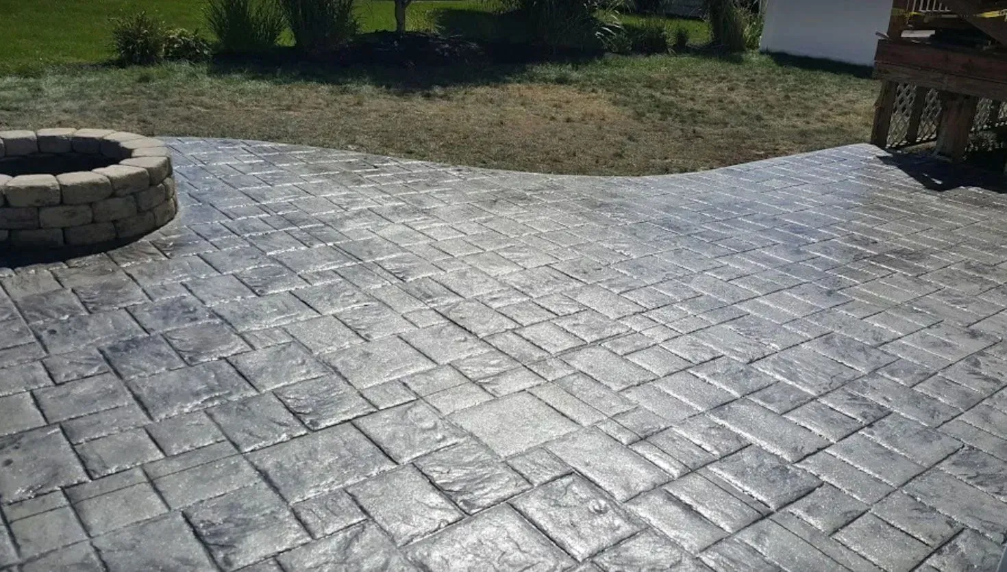 A grey, stamped concrete patio with a rectangular brick pattern and a circular stone fire pit in a backyard setting.