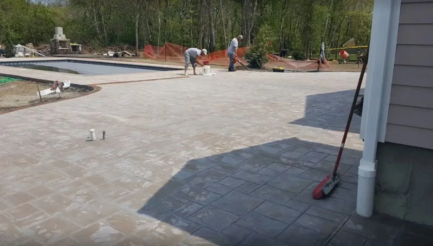Two people work on a stone patio installation outside a house, with a pool in the background.