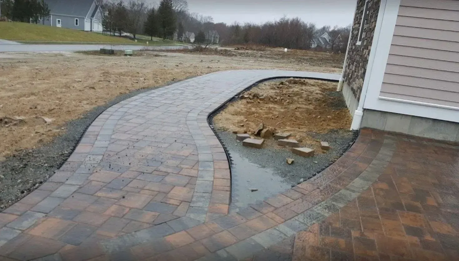 A curved stone paver walkway leads toward the corner of a house, alongside a dirt garden bed and paved patio area.