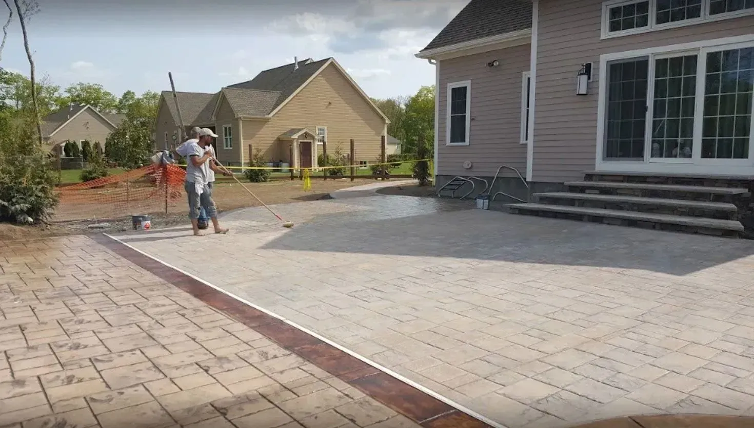 A worker uses a long-handled tool to sweep a newly installed light-colored paver patio beside a tan house.