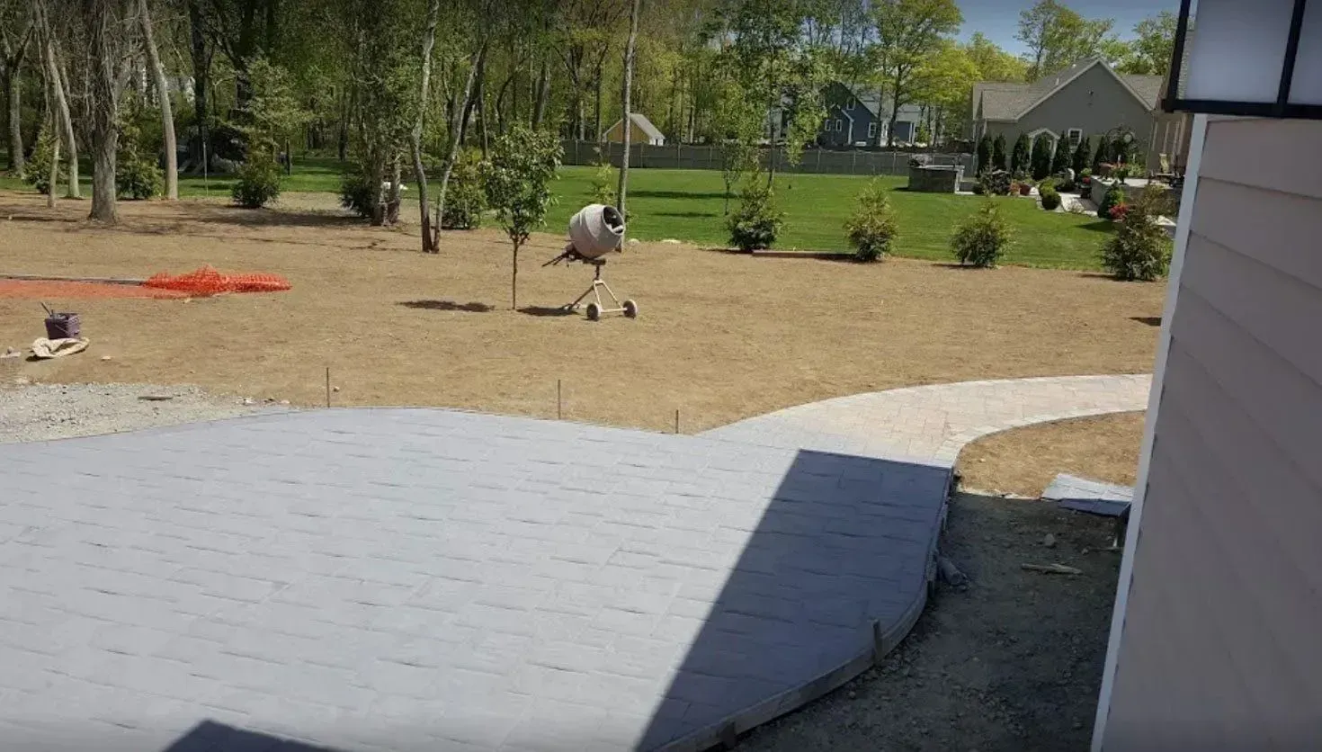 A gray stamped concrete patio under construction extends into a large, sunny yard with grass and trees.