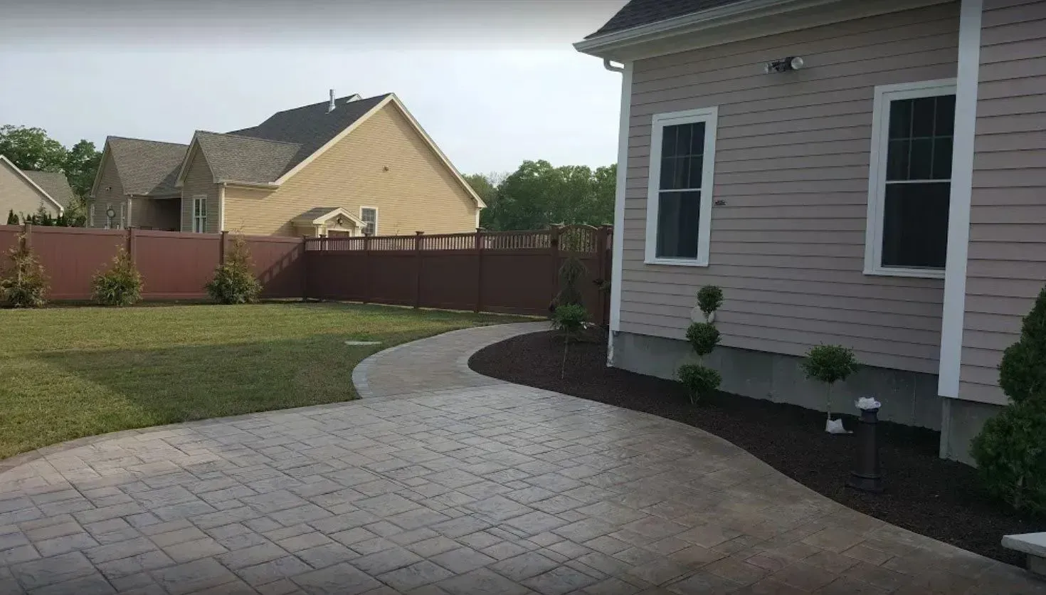 A patio with paver stones leads to a backyard path alongside a light-colored house and a brown fence.