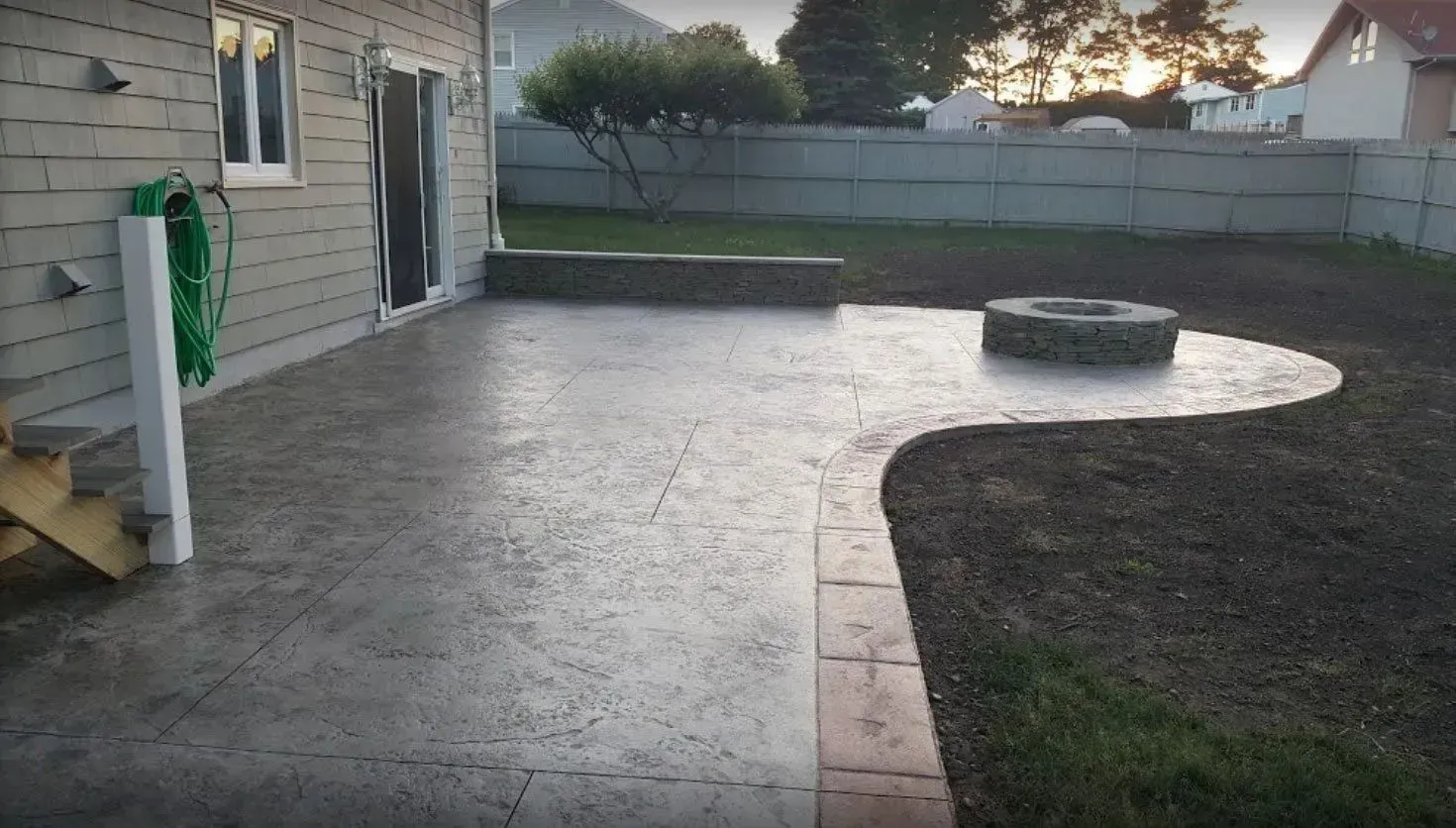 A stamped concrete patio featuring a built-in fire pit and a stone wall bordering a backyard at sunset.