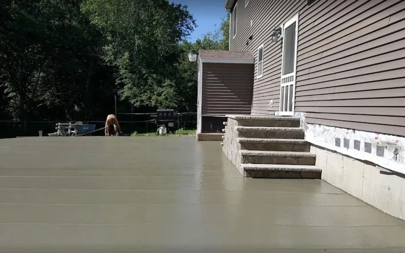 A freshly poured concrete patio next to a house with brown siding and a small set of concrete steps.