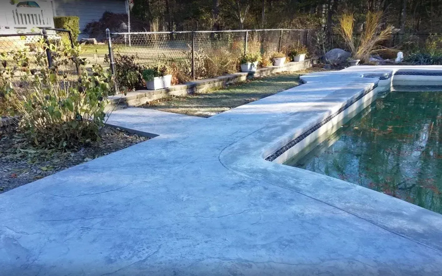 A concrete pool deck surrounding the edge of a backyard swimming pool, with a fence and landscaping in the background.