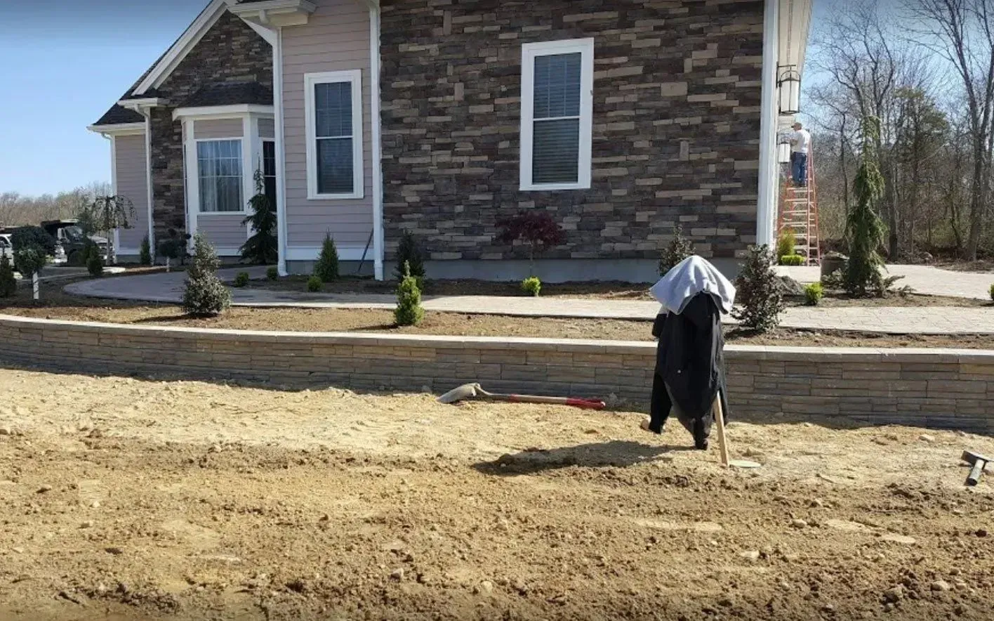 A person wearing a head covering stands near a stone retaining wall in front of a house under construction.