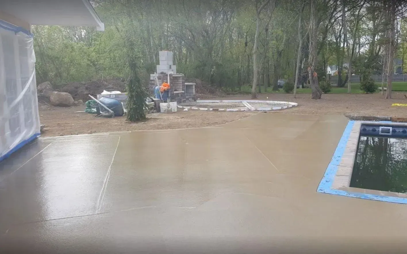 A backyard under construction with fresh concrete, a pool edge, and an outdoor fireplace area in a wooded setting.