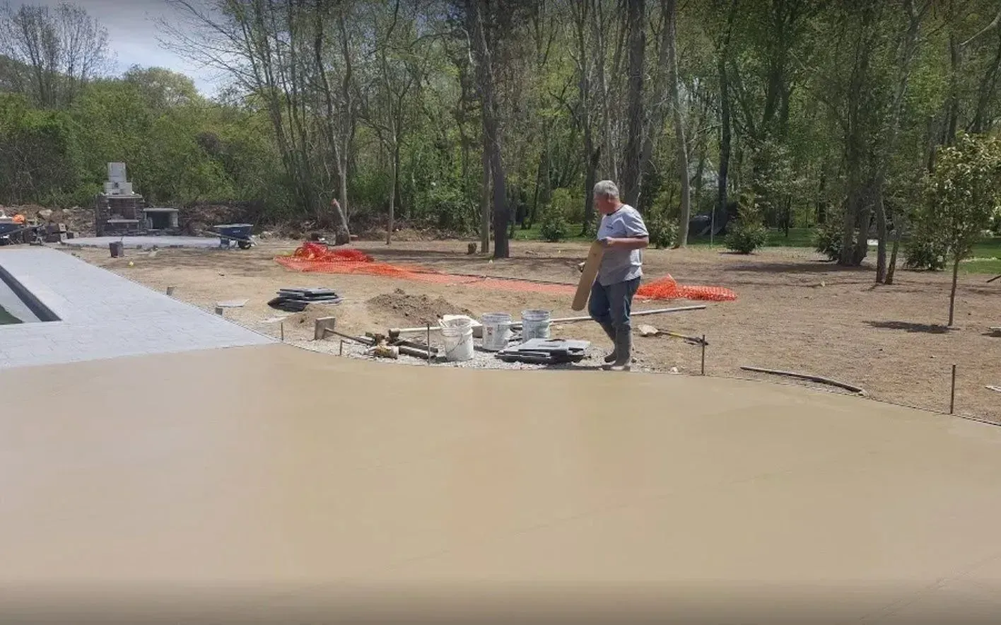 A worker holding a wooden board stands on a newly poured concrete patio in a yard under construction.
