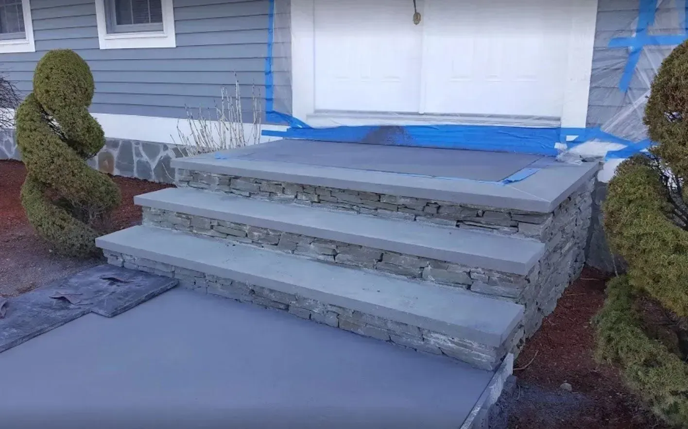 A set of stone steps with concrete treads leading to a house entrance, flanked by spiral topiary shrubs.