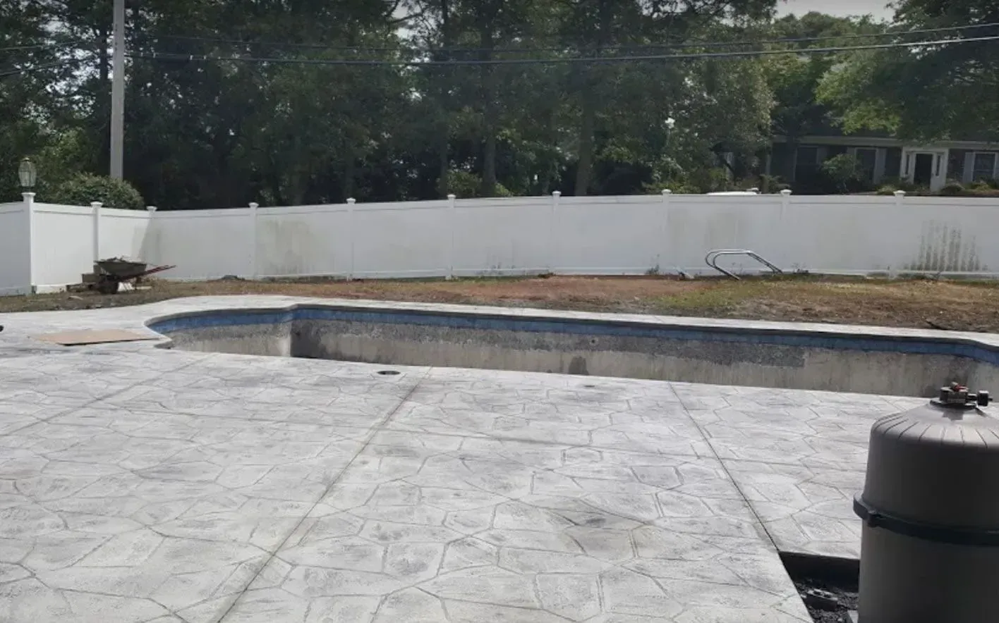 An empty swimming pool with a stone-textured concrete deck, surrounded by a white vinyl fence and trees in a backyard.