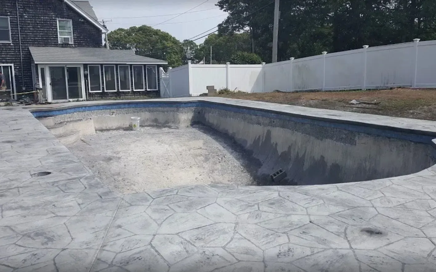 An empty, grey-tiled swimming pool under construction, surrounded by stamped concrete decking and a white privacy fence.