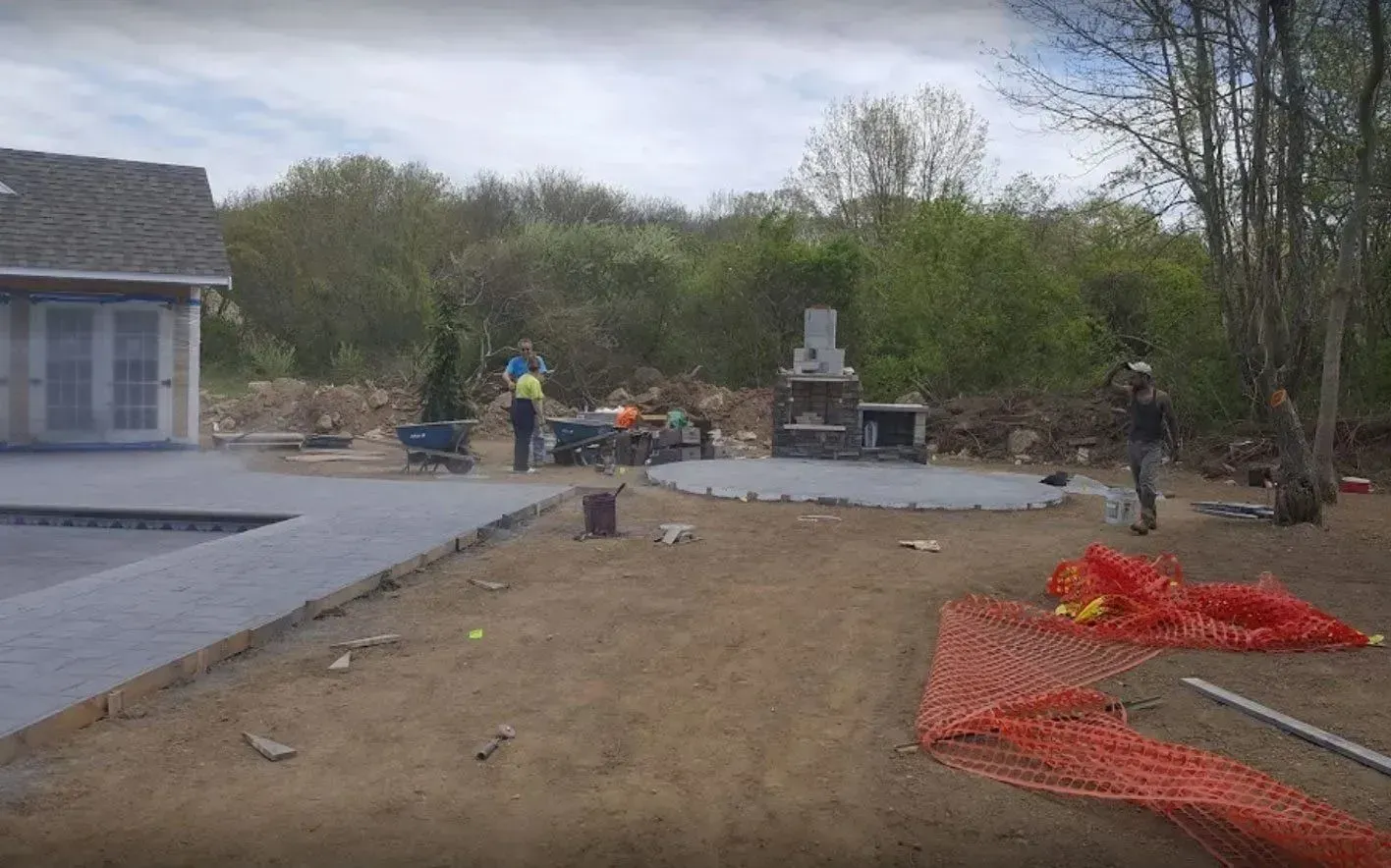 Workers installing a stone patio and fireplace in a backyard next to a pool under construction.
