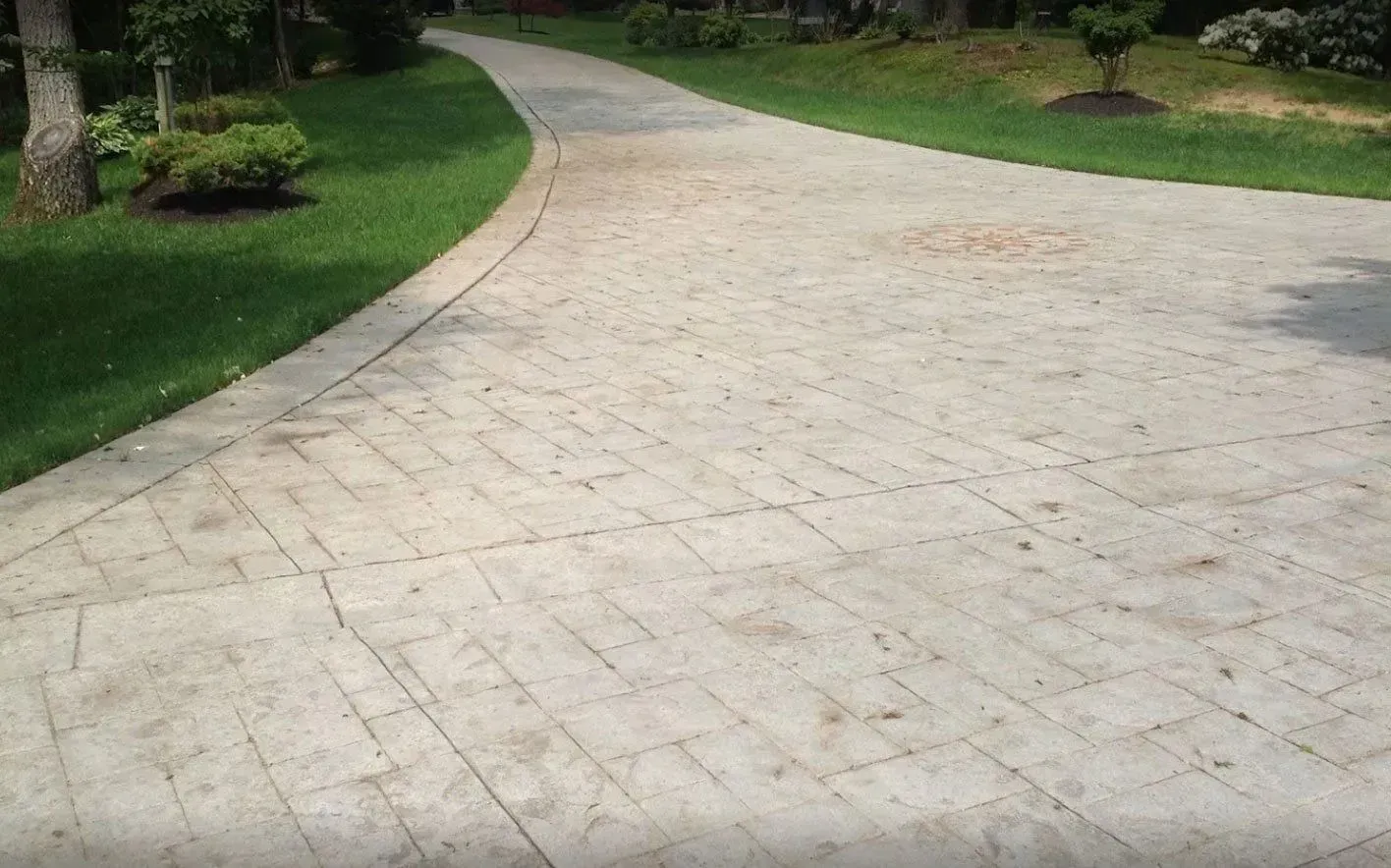 A beige stamped concrete driveway with a grid pattern, curving past a green lawn with shrubs and trees.