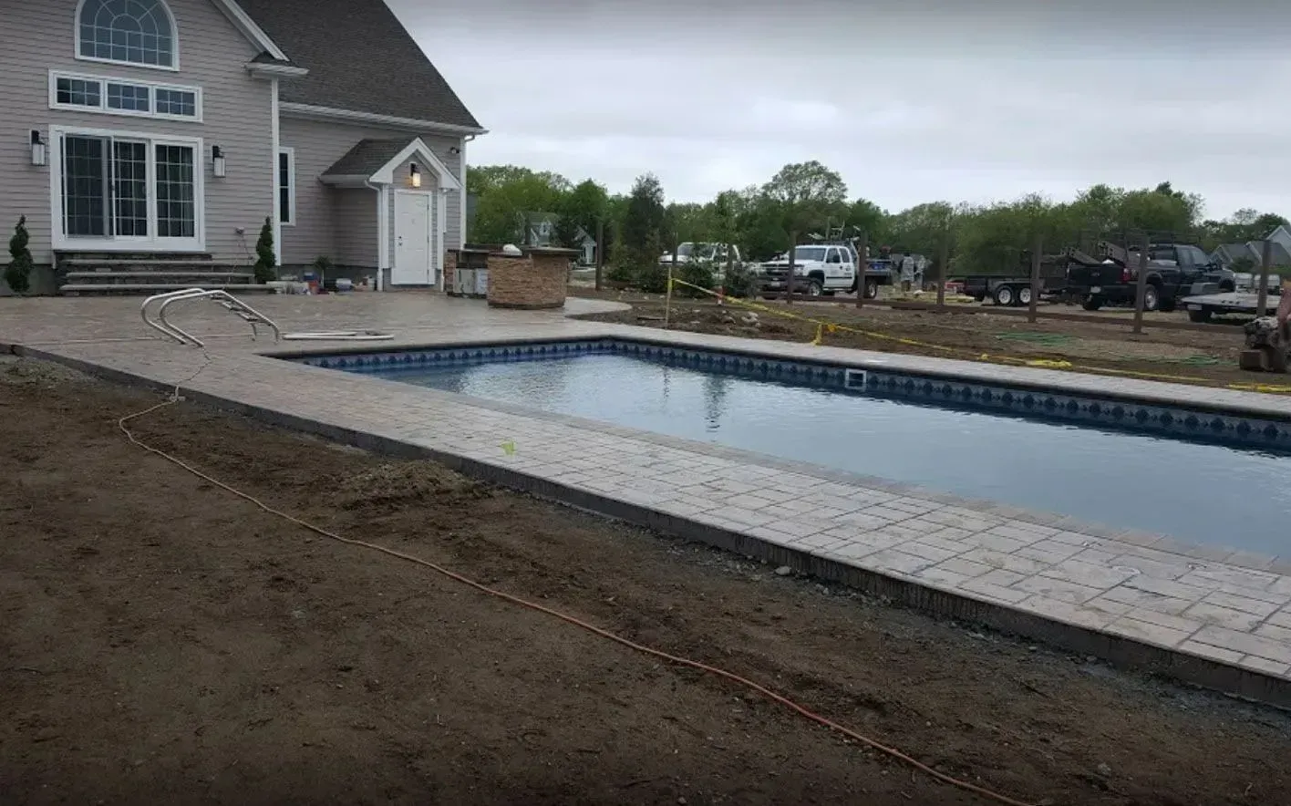 A rectangular backyard pool with stone patio edging next to a gray house, surrounded by unfinished landscaping.