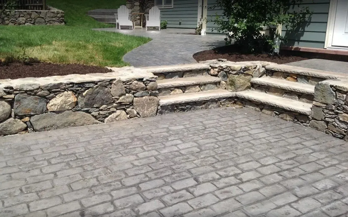 A stone patio with a low rock retaining wall and steps leading up to a paved walkway and a house.