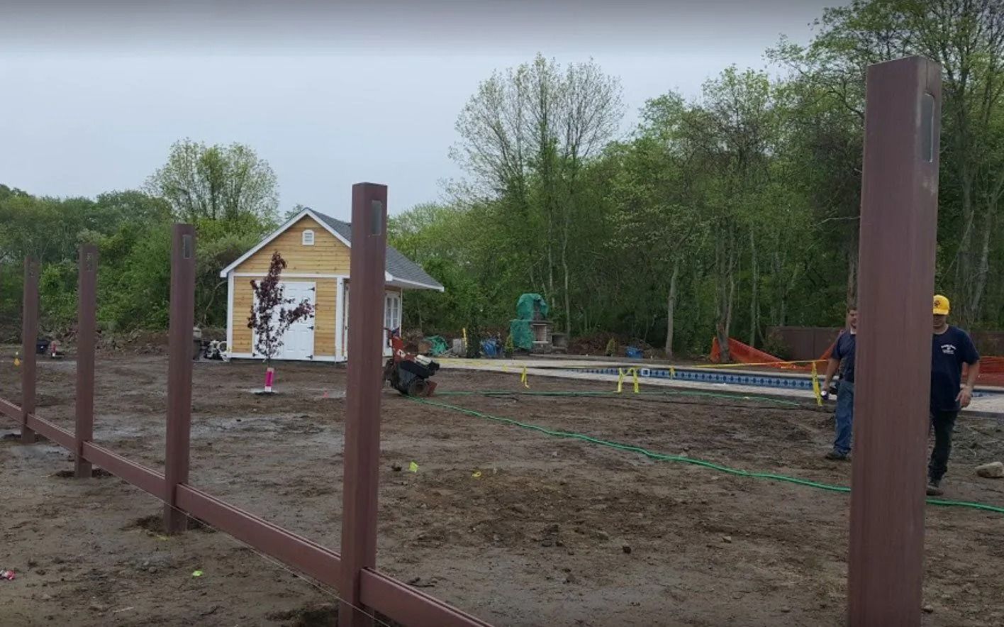 Two people stand on a muddy construction site near brown fence posts and a small yellow building.