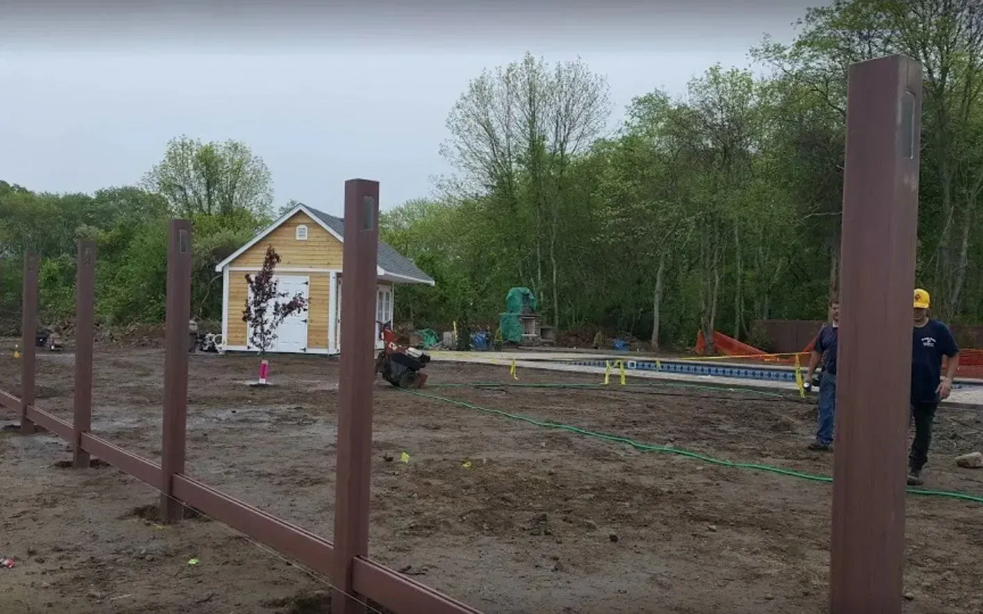 Two people work on a construction site with several tall brown posts installed in a muddy, grassy yard near a small shed.