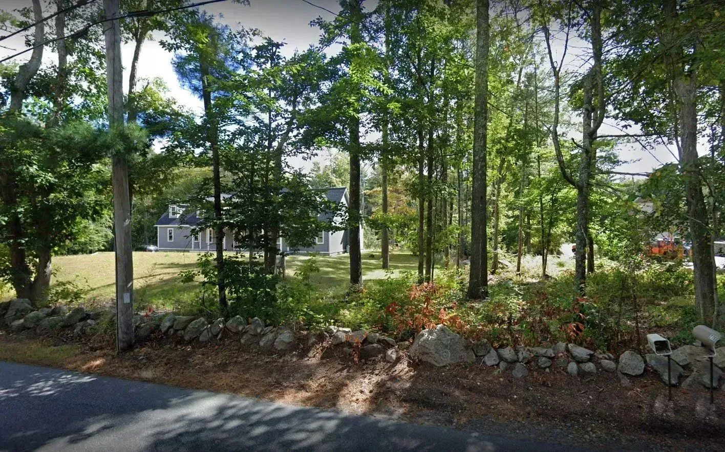 A gray, two-story house sits set back behind trees and a stone wall along a paved road.