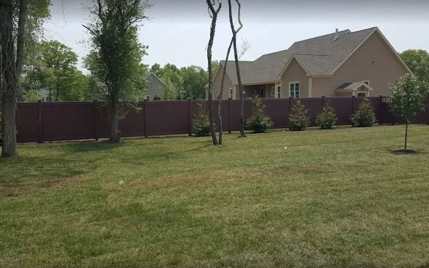 A grassy backyard with a tall, dark-red privacy fence and several young trees in front of a tan brick house.