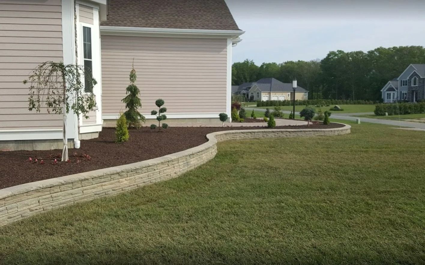 A tan stone retaining wall curves along a landscaped yard with dark mulch and small decorative trees in front of a house.