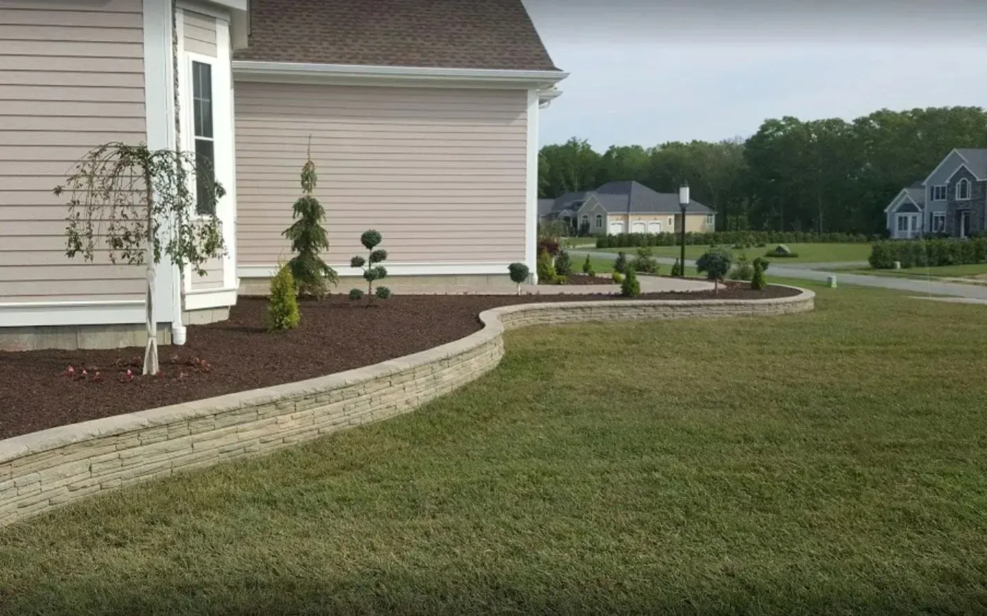 A beige house exterior with a curved stone retaining wall surrounding a garden bed with mulch and small trees.