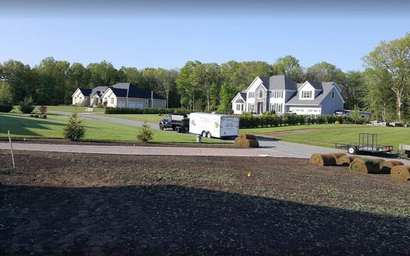 A bright, sunny day shows a suburban yard under construction, with dark soil, stacks of sod, and houses in the background.