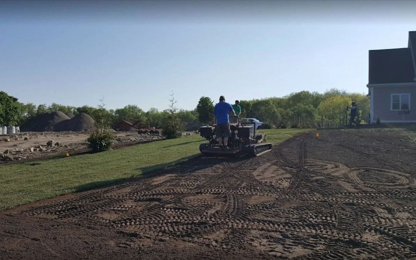 A person operates a tracked skid steer to grade a dirt yard next to a house on a sunny day.