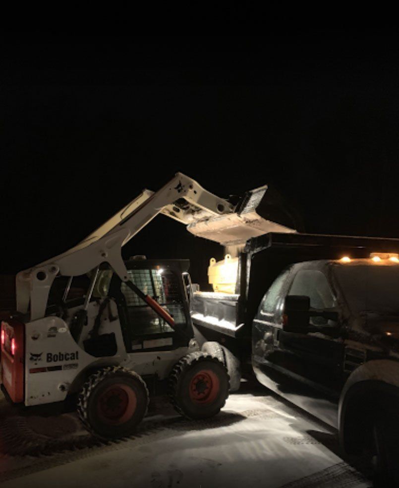 A white Bobcat skid-steer loader dumps material into the back of a black dump truck at night.