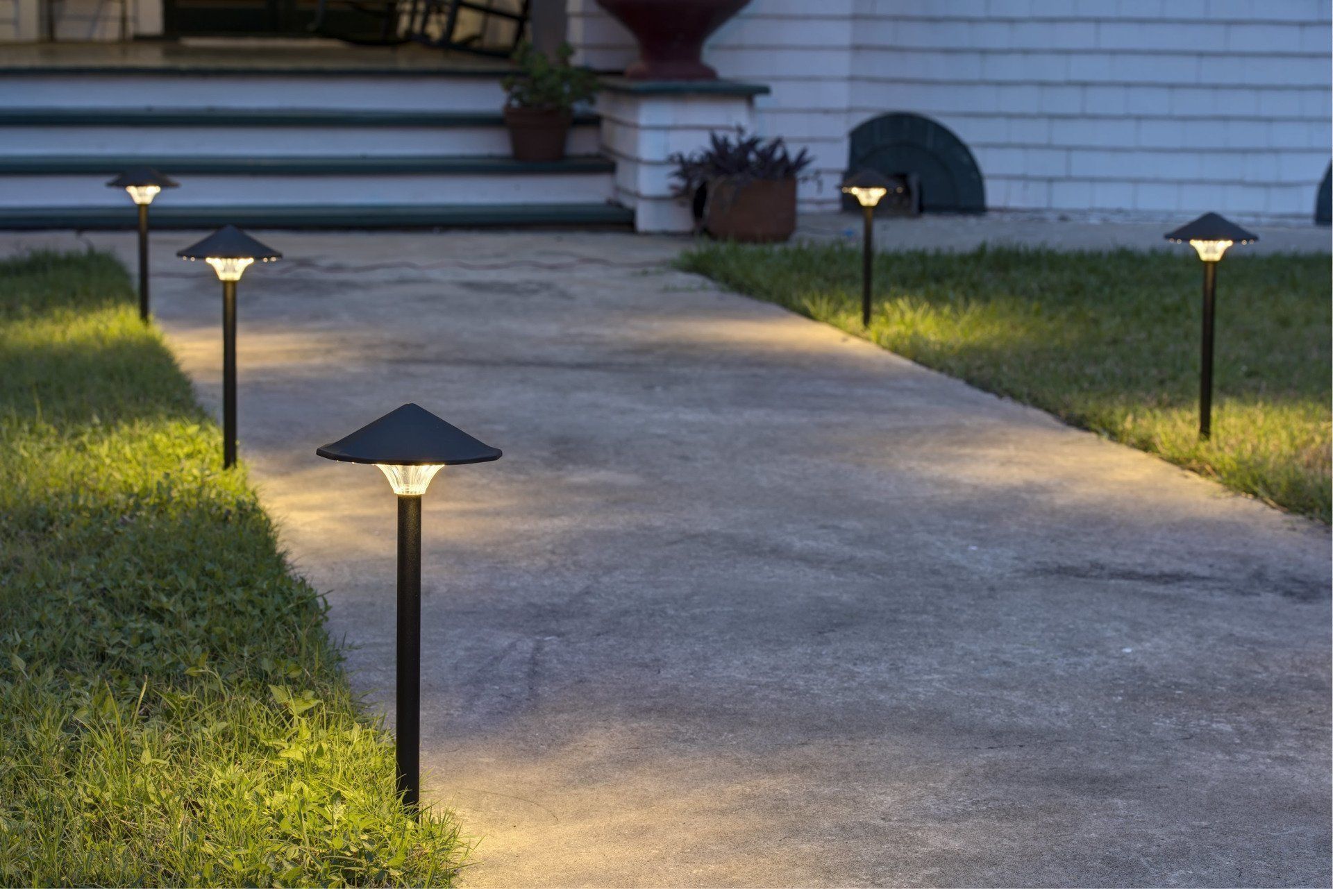 A paved garden path illuminated at night by several modern, black stake lights with conical shades along the grass edges.
