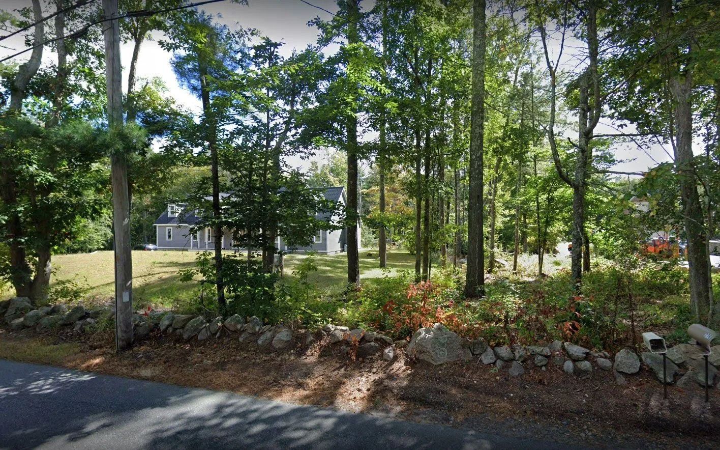 A grey house stands on a grassy lot behind a stone wall, framed by tall trees and viewed from a paved road.