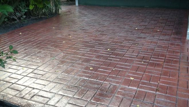 A wet, reddish-brown driveway featuring a herringbone brick pattern, with a dark green garage door in the background.