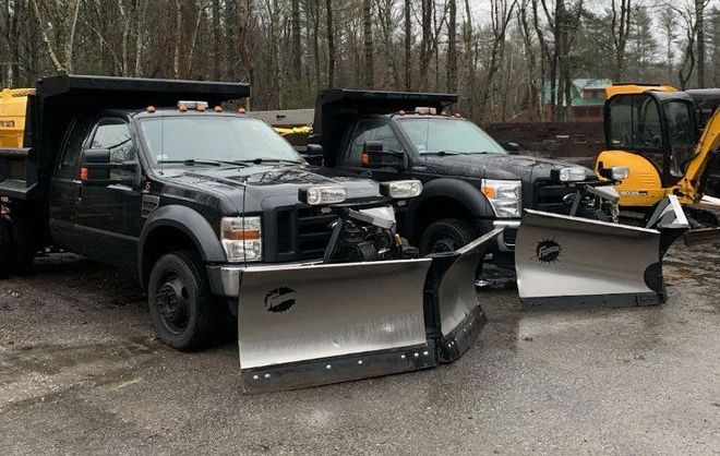 Two black pickup trucks equipped with snow plows parked on an asphalt lot with trees in the background.