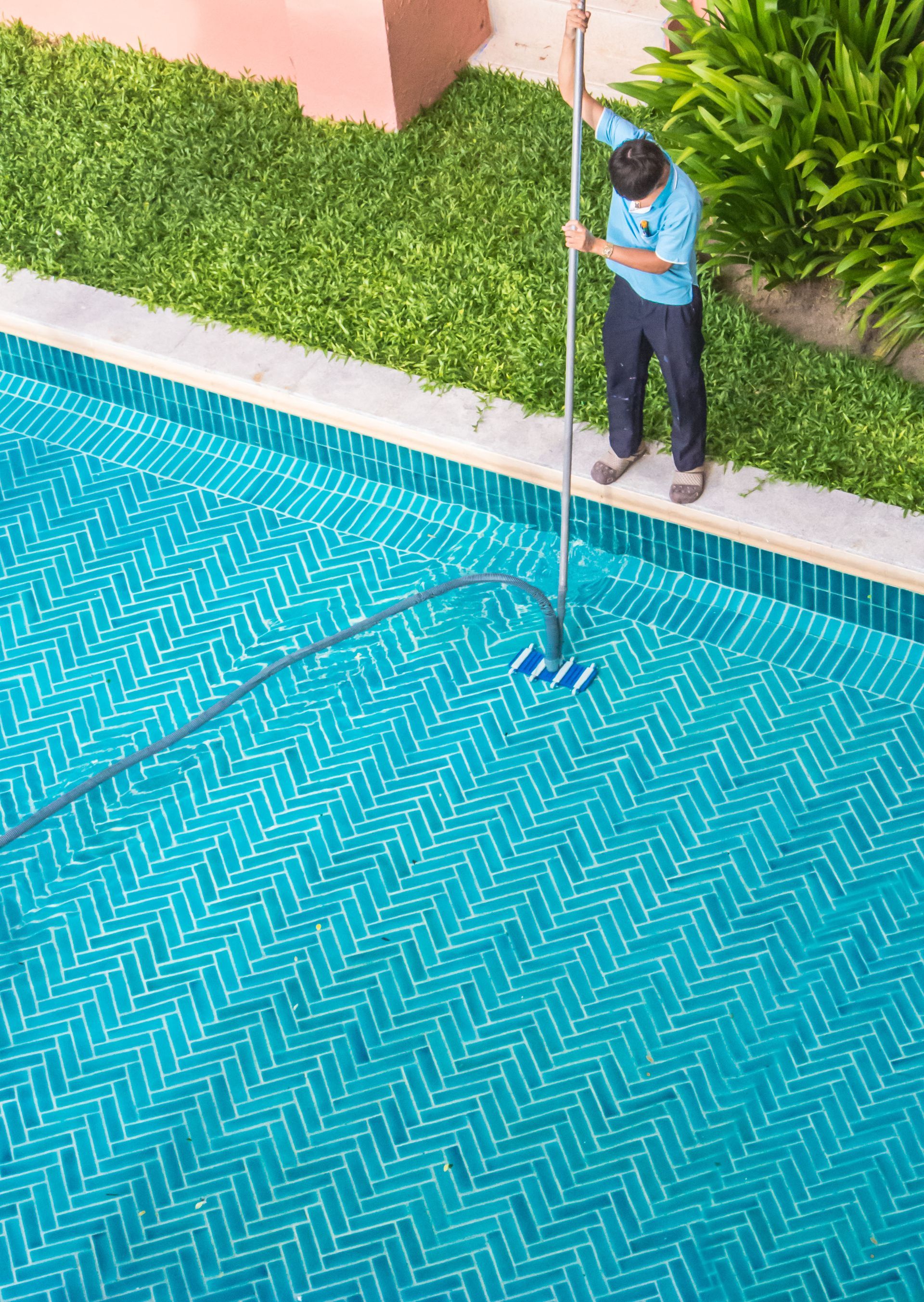 Person cleaning a turquoise tiled swimming pool with a long-handled brush. Green grass and plants are nearby.