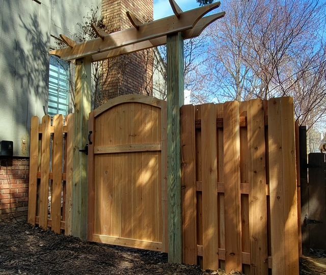 A light brown wooden fence with an arched gate, framed by two tall posts supporting a decorative horizontal pergola top.